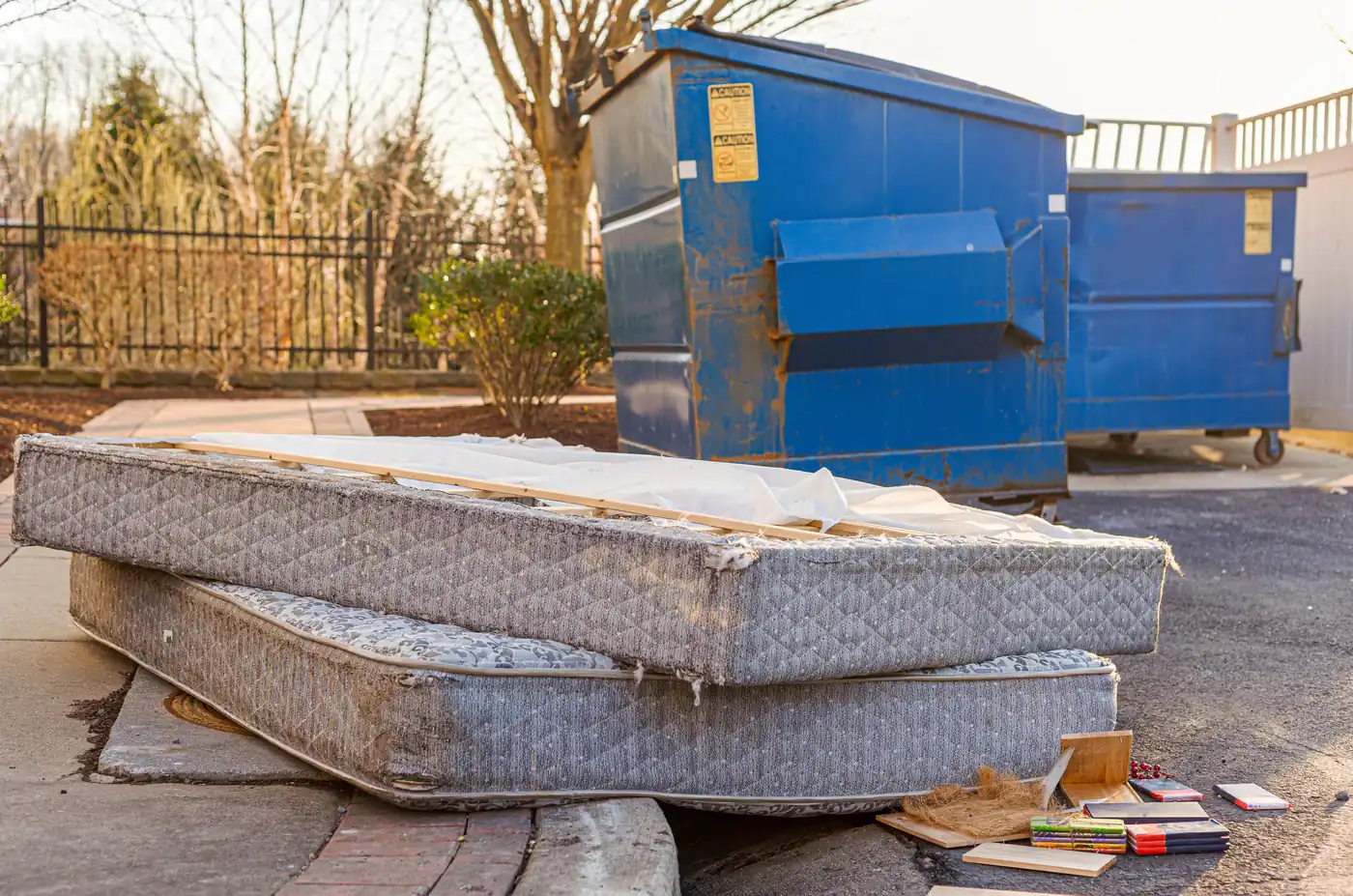 Discarded mattress and box spring stacked at the curb beside apartment dumpsters